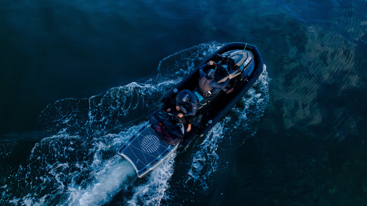 Overhead drone perspective of a black rigid inflatable boat cutting through deep blue ocean water, with two wetsuit-clad spearfishers aboard and a Waihana-logo swim platform at the stern.